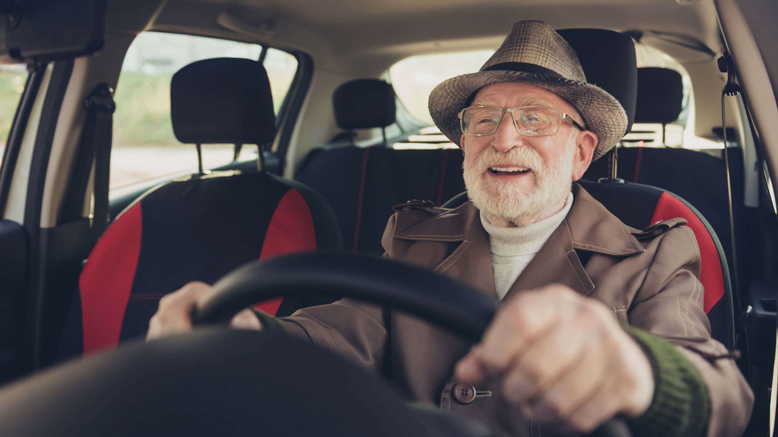 Ein älterer Mann mit Brille und Bart sitzt am Lenkrad und lächelt.